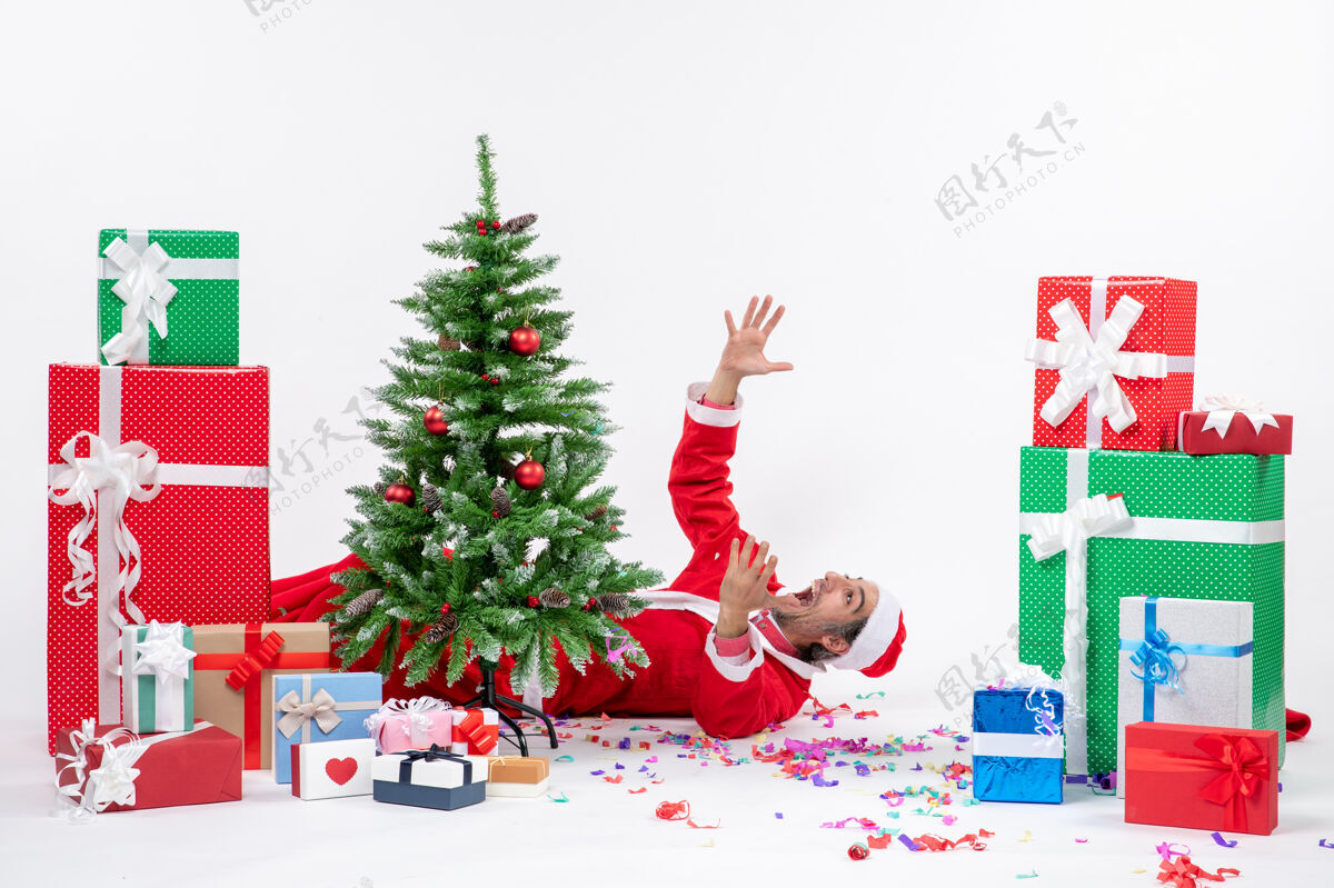 圣诞老人节日气氛与年轻紧张的圣诞老人躺在圣诞树后靠近礼物白色背景冬青圣诞老人说谎