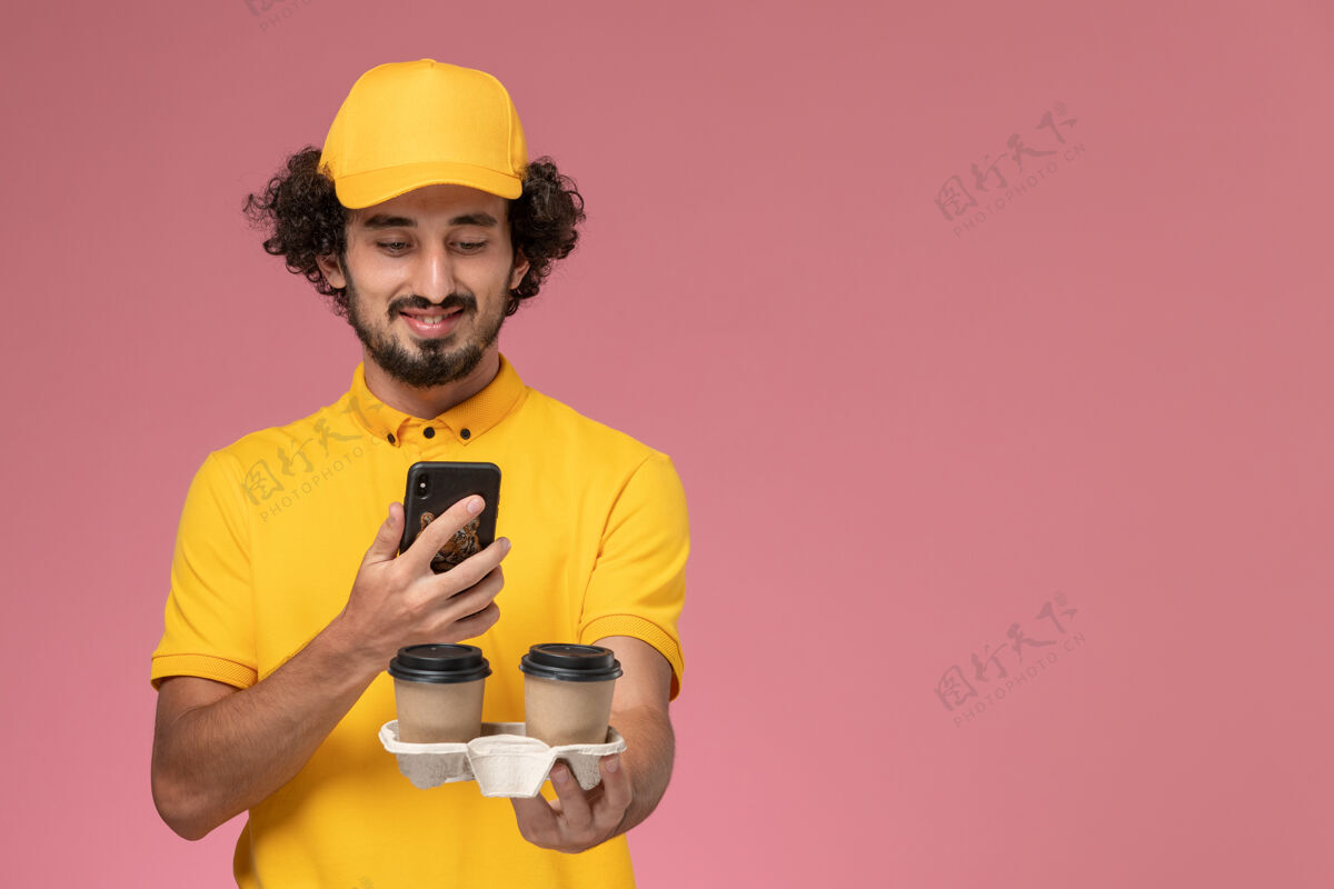 工人正面图穿着黄色制服和披风的男信使拿着棕色的咖啡杯在粉红色的墙上拍照制服粉色男