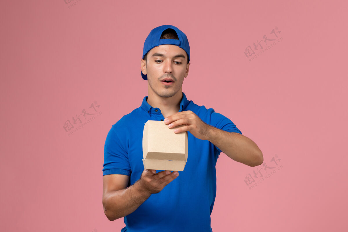 包裹正面图：身穿蓝色制服披肩的年轻男性快递员在粉红色的墙上拿着小包裹成人快递员正面