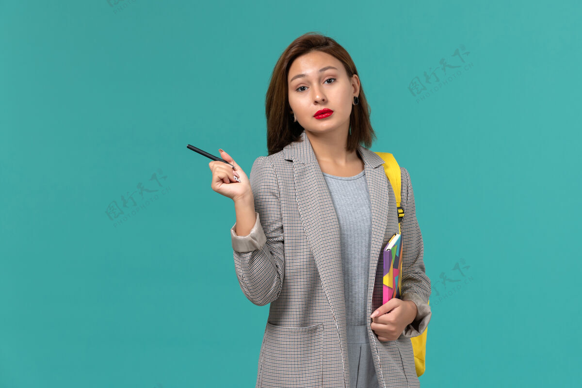 大学穿着灰色夹克 背着黄色背包 拿着文案的女学生的正面图 蓝色的墙上有钢笔学生穿戴举行