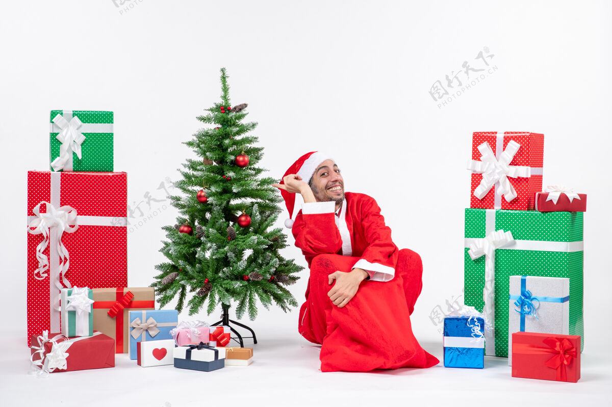 装饰微笑着兴奋的年轻人打扮成圣诞老人与礼物和装饰圣诞树坐在地上的白色背景微笑礼物地