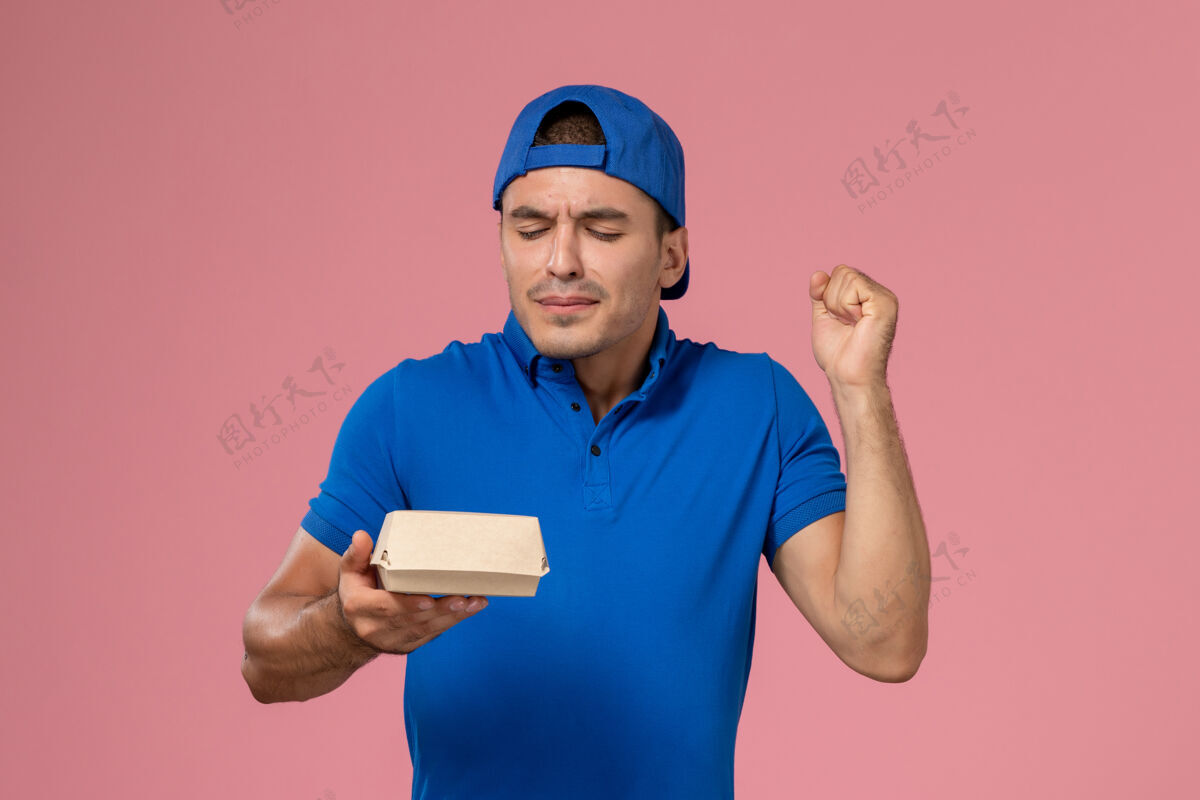男性正面图身穿蓝色制服披肩的年轻男性快递员拿着小快递食品包站在浅粉色的墙上送货年轻男性快递食物