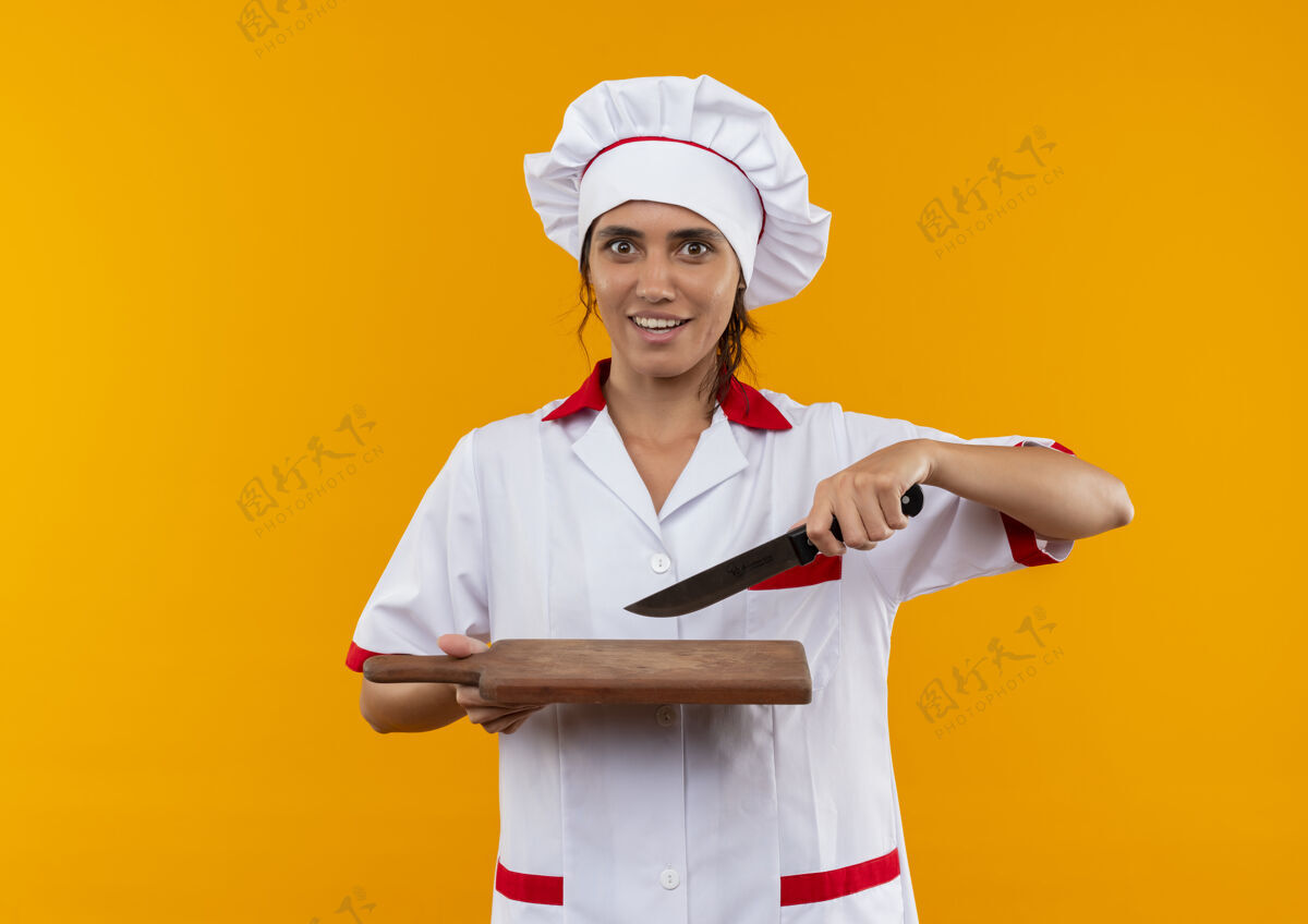 厨师令人印象深刻的年轻女厨师穿着厨师制服拿着刀和砧板在隔离的黄色墙壁与复制空间刀穿年轻